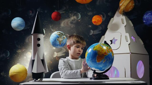 A boy in a striped sweater sits at a table and spins a globe. Next to him is a model rocket, and in the background, there is a cardboard model of a spaceship and hanging planets. The scene resembles a