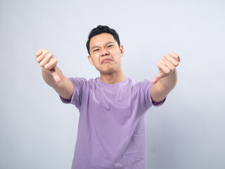 Young Asian man in a lavender t-shirt showing a disapproving expression and giving a thumbs-down gesture. Studio shot on a plain background, highlighting his displeased expression