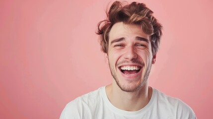 A young man with brown hair and a beard laughs in front of a pink background