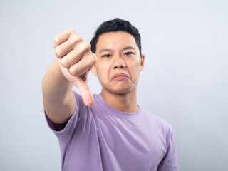 Young Asian man in a lavender t-shirt showing a disapproving expression and giving a thumbs-down gesture. Studio shot on a plain background, highlighting his displeased expression