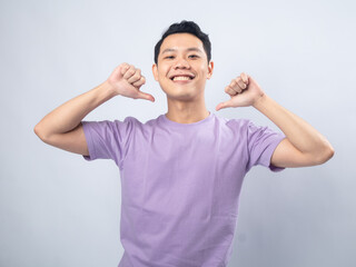 Fototapeta premium Young Asian man in a light purple shirt smiling confidently while pointing at himself with both thumbs, standing against a plain light grey background. 
