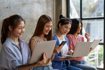 Four young businesswomen are working from home, sitting on a bench by the window and using a laptop computer, a digital tablet and a mobile phone