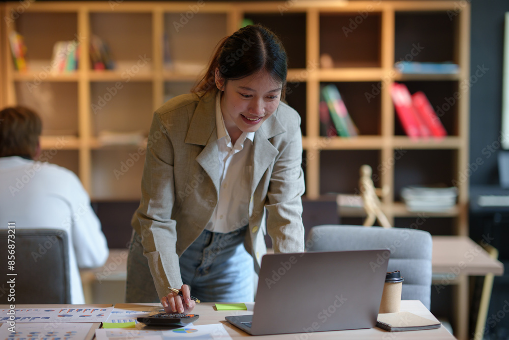 Poster young asian businesswoman leaning over desk and using calculator while working on laptop computer in