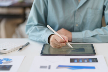 Businesswoman is using a stylus pen to analyze financial data on a tablet. She is working at a desk with charts and graphs spread out in front of her