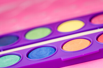 Close up of a watercolor paint palette with a paintbrush, vibrant colors on a pink background.