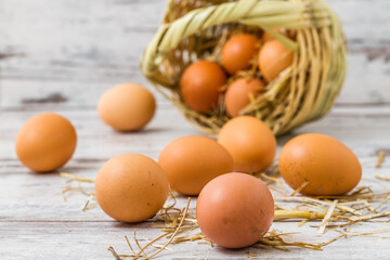 Fresh brown eggs, some speckled, scattered on a white wooden surface and inside a tipped over woven basket, surrounded by strands of straw.