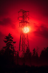 Naklejka premium Dramatic sunset behind an electricity pylon with dark trees, creating a striking silhouette against a vivid red and orange sky.