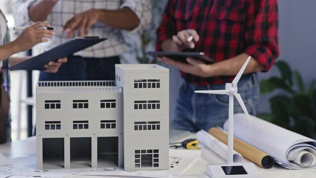 Architects analyzing blueprints and building model with wind turbine, planning sustainable designs in a modern office. Team collaboration for eco-friendly construction