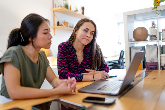 Asian woman and Aboriginal woman sitting together at a desk in office looking at laptop