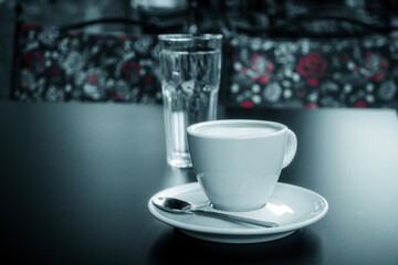 A white cup of coffee and a glass of water on a black table in a sidewalk cafe. Floral pattern on blurred chairs in the background