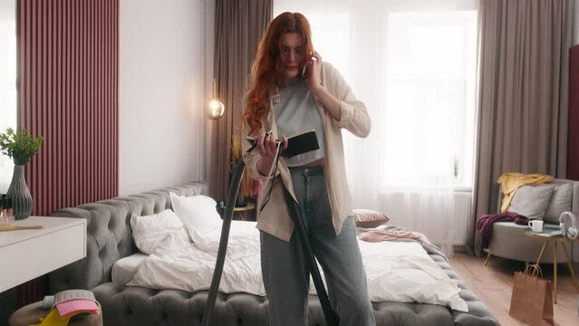 Busy multitasking Caucasian woman girl housewife businesswoman work from home deadline stressed cleaning mess bedroom with vacuum cleaner anxious annoyed female talk mobile phone housekeeping chores