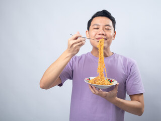Young Asian man in a lavender t-shirt enthusiastically eating instant noodles from a bowl with a...