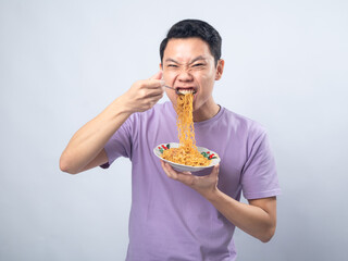 Young Asian man in a lavender t-shirt enthusiastically eating instant noodles from a bowl with a fork. Studio shot on a plain background, highlighting his joyful expression and casual fashion © Daniel