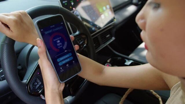 Close up of young Asian woman monitoring status of battery on application on mobile phone while sitting inside electric car