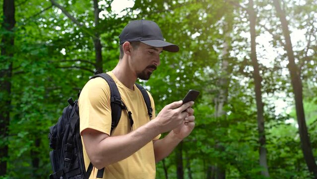 Face man with beard and backpack in forest uses phone to touch the screen looking for connection signal gps application orientation tourist travel hike lost sun communication telephone
