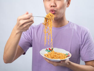 Young Asian man in a lavender t-shirt enthusiastically eating instant noodles from a bowl with a...