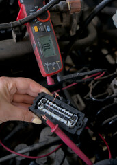 An auto electrician using a measuring device to check the voltage in a vehicle's electrical circuit, diagnosing wiring issues for repair. Auto service.