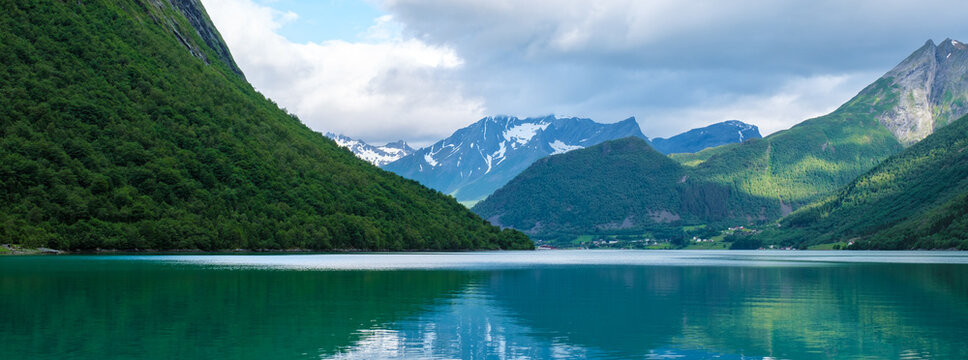 A picturesque view of a fjord in Norway, with lush green mountains framing the tranquil water. The sky is partly cloudy, creating a soft, natural light. Norangsfjorden Norangdal Urke Norway