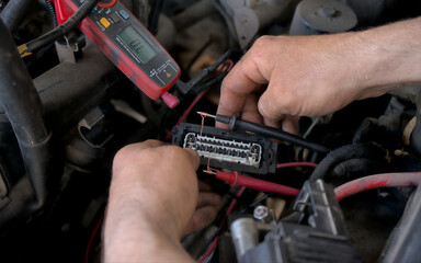 An auto electrician using a measuring device to check the voltage in a vehicle's electrical circuit, diagnosing wiring issues for repair. Auto service.
