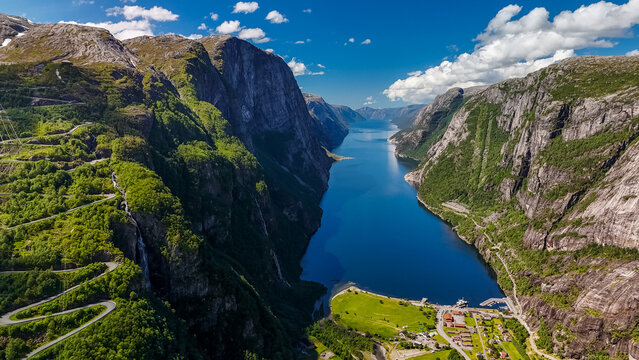 A scenic aerial view of a winding road carved into the steep cliffs of a Norwegian fjord, with a picturesque village nestled at the base. Lysebotn, Lysefjorden, Norway