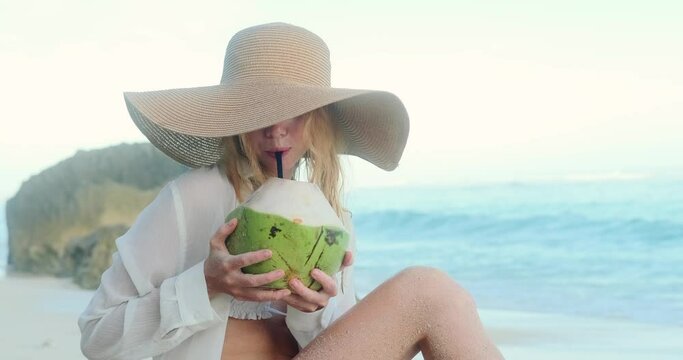 A young attractive woman in a straw hat and white bikini sits on the sand on the beach near the ocean and drinks a fresh coconut through a straw. Summer vacation on the tropical island of Bali.