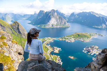Naklejka premium A woman sits on a rocky cliff overlooking a stunning fjord landscape in Norway, enjoying the breathtaking views of the mountains and the sparkling blue waters. Reinebringen, Lofoten, Norway