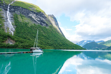 A white sailboat is docked at a wooden pier, nestled in a picturesque Norwegian fjord surrounded by...
