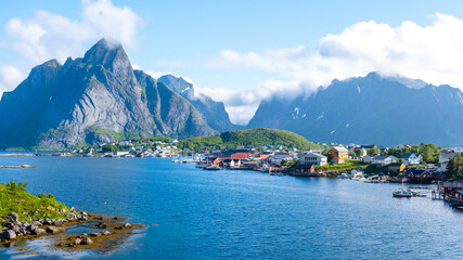 Fototapeta premium Reine, Lofoten, Norway, village nestled between towering mountains in Norway. The clear blue waters of the fjord create a stunning contrast with the lush greenery and the traditional red houses.