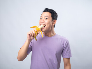 Fototapeta premium Young Asian man in a lavender t-shirt smiling while eating a peeled banana. Studio shot on a plain background, highlighting his casual fashion and cheerful expression.