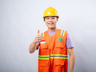An Asian male construction worker, wearing an orange safety vest and a yellow hard hat, gives a double thumbs-up gesture while smiling. The plain white background highlights the worker 