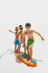 Three boys in swim trunks and sunglasses balancing, as surfing on inflatable board against white...