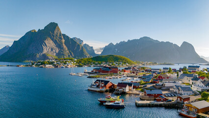 A picturesque coastal village in Norway, surrounded by towering mountains, showcasing red-roofed houses and boats in the calm waters of a fjord. Reine, Lofoten, Norway