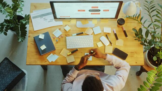 Black businessman using computer, writing sticky notes and attaching them to desk during workday in the ofice. Time lapse, directly above view
