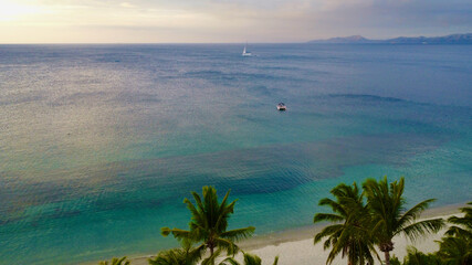 Aerial view of the warm sea and the shore of a tropical island. Top view of the coast of a tropical island, beach and palm trees, smooth surface of a calm sea and clouds above the horizon.