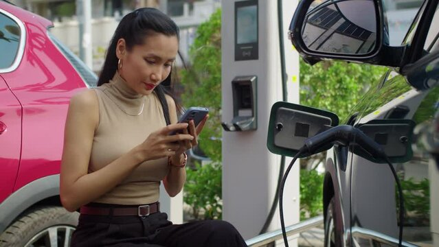 Medium long shot of young Asian businesswoman messaging on mobile phone when waiting for her electric car to charge at city station