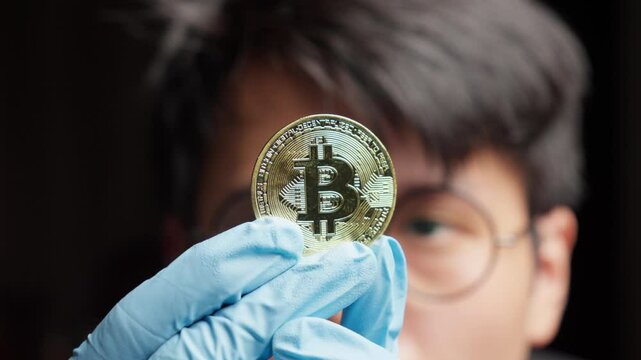Asian man wearing gloves holds a coin representing BTC, analyzing technology