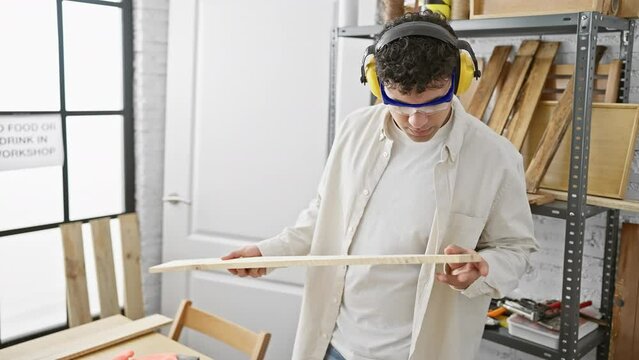A focused young man with safety glasses and earmuffs examines a piece of wood in a bright carpentry workshop