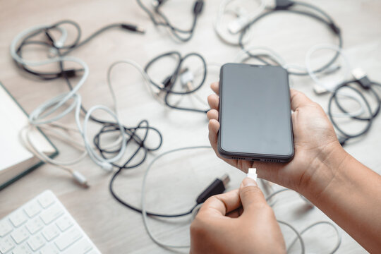 Closeup hands of a woman holds a smartphone, going to charge battery with an incompatible USB wire, with many electric USB cords on the table, confused plugs and sockets