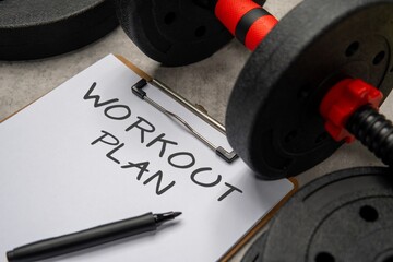 A close-up image of a clipboard with a WORKOUT PLAN written on it next to a black dumbbell and weight plates. Sport and fitness concept.