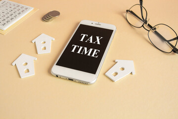 Tax Time and house symbol. Concept words Tax Time written on the mobile phone screen. Beautiful beige background. Glasses, silver coins, miniature houses. Business and tax time house concept.