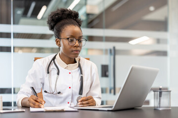 Doctor diligently working on laptop and writing notes in a modern medical office. Professional healthcare provider wearing a stethoscope, demonstrating concentration and dedication to her work.