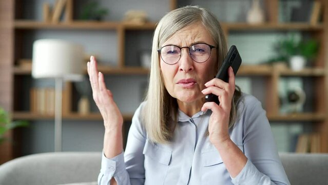 Angry excited senior female talking arguing over phone while sitting on sofa in living room at home. Frustrated elderly woman with gray hair complains about problems using a smartphone. Close up