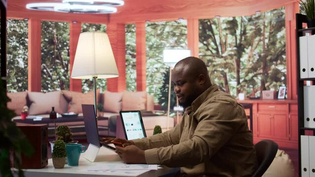 Young self employed businessman does online shopping with his card, making electronic banking payments at his home office. Freelancer does remote work and purchases things on laptop. Camera B.