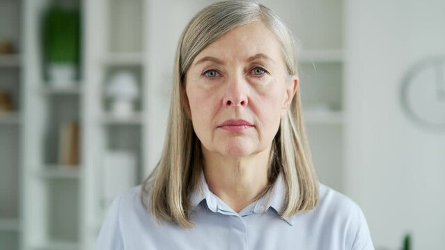 Portrait of serious senior gray haired businesswoman standing in living room at home. Confident middle aged woman looking at the camera. Headshot of a focused female entrepreneur indoors. Close up