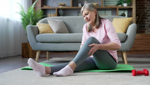 Senior woman suffers from leg pain, calf muscle spasm during a gymnastics workout sitting on floor on a mat in the living room at home. Elderly woman massaging leg calf muscle cramps tendon at calves