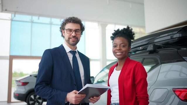 Mixed-raced people glancing at camera at same time. Attractive man in business costume holding folder and standing next to pretty woman. Consultant and client happy about successful deal.