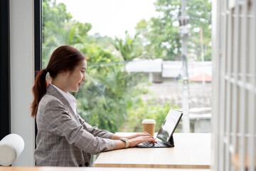 An attractive, thoughtful Asian businesswoman is working remotely from a cafe, using her digital tablet, gazing at the view outside, and thinking about her new ideas.