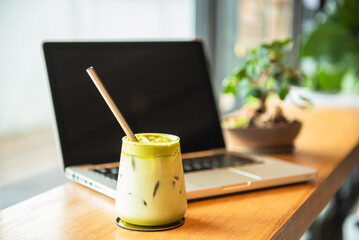 Iced matcha latte glass with metal straw Placed in front of an open laptop on a wooden table. There is a small potted plant. The back is blurry. Reflects the comfortable atmosphere in a coffee shop.