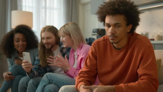 African American sad offended frustrated man guy male at home separated ignoring looking at group of multiracial Caucasian multiethnic friends women men mobile phone using addicted laughing gossiping