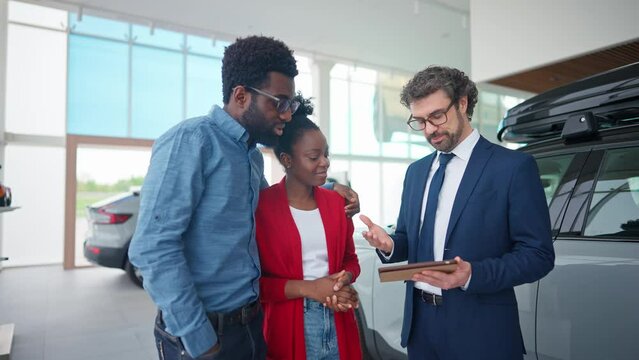 Handsome auto dealer helping mixed-raced customers to choose car that fits their needs and budget. Man in business costume holding tablet device and talking with married couple in vehicle centre.